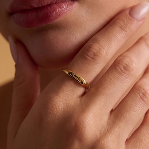 Könnte beinhalten: Ein polierter goldfarbener Ring mit einem flachen, rechteckigen Mittelteil, in den der Name Sophia in Schreibschrift eingraviert ist. Der Ring hat eine glatte, reflektierende Metalloberfläche und ein schmales, abgerundetes Banddesign.
