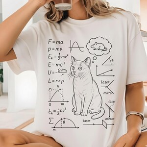 Puede incluir: Camiseta blanca con una ilustración en blanco y negro de un gato, ecuaciones científicas y diagramas. Una burbuja de pensamiento sobre el gato muestra un ratón. La camiseta tiene cuello redondo.