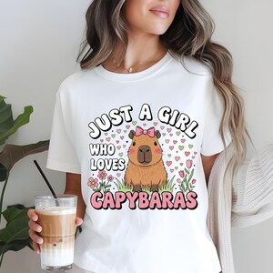 May include: White t-shirt with the text "JUST A GIRL WHO LOVES CAPYBARAS" in a playful font. The design features a cartoon capybara with a pink bow, surrounded by hearts and flowers. The shirt is a casual, everyday wear item.