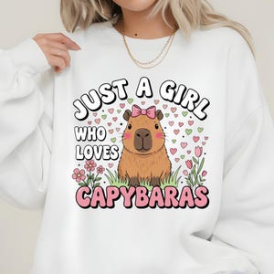 May include: White sweatshirt with a graphic of a capybara wearing a pink bow, surrounded by hearts and flowers. The text reads "JUST A GIRL WHO LOVES CAPYBARAS" in a playful font. A cute and cozy design.