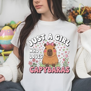 May include: White t-shirt with the text "JUST A GIRL WHO LOVES CAPYBARAS" in a playful font. The design features a cartoon capybara with a pink bow, surrounded by hearts and flowers. A cute and whimsical design.