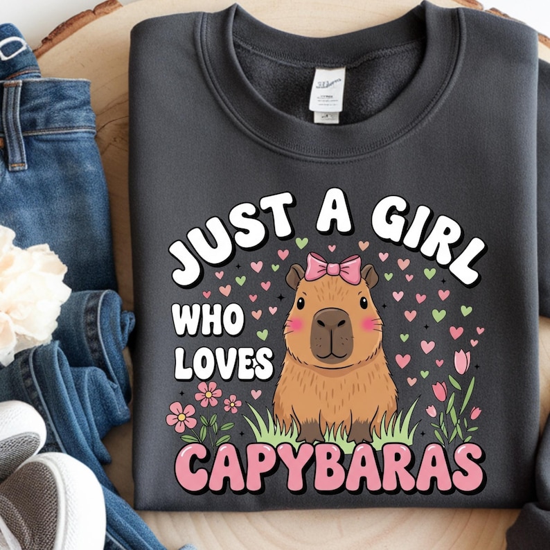 May include: Dark gray sweatshirt with the text "JUST A GIRL WHO LOVES CAPYBARAS" in white and pink. The design features a cartoon capybara with a pink bow, surrounded by hearts and flowers.