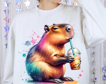 May include: White sweatshirt featuring a colorful illustration of a capybara drinking boba tea. The capybara is depicted with rainbow-colored fur and is holding a clear plastic cup with a blue straw. Watercolor paint splatters surround the capybara.