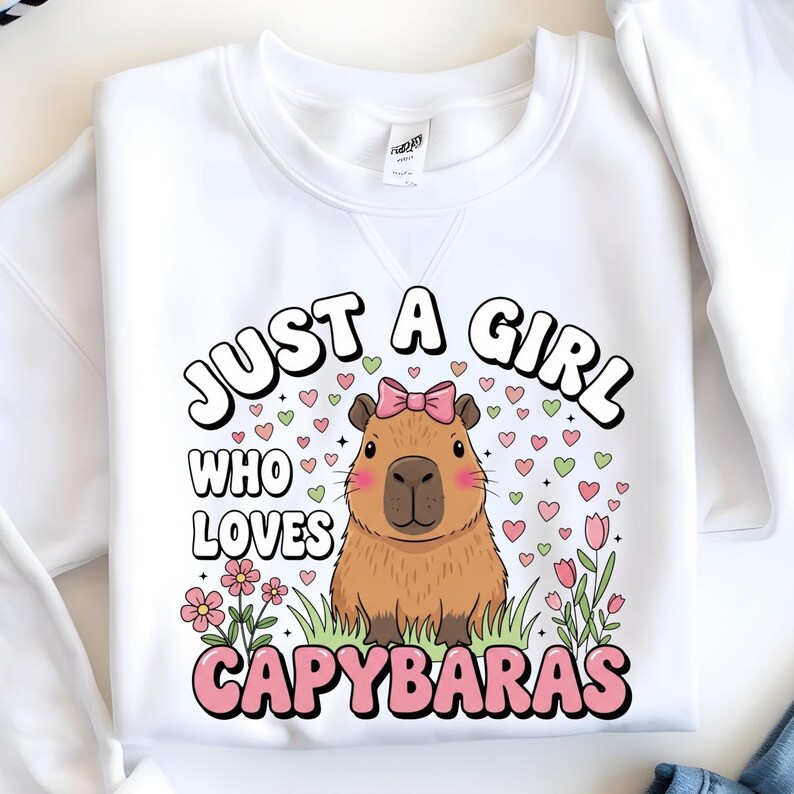 May include: White sweatshirt featuring a cartoon capybara with a pink bow, surrounded by hearts and flowers. Text reads "JUST A GIRL WHO LOVES CAPYBARAS" in a playful font. A cozy and cute design.