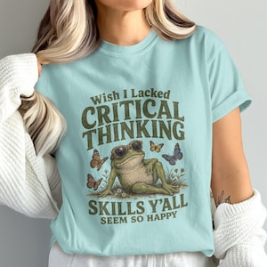 May include: A light blue t-shirt featuring a graphic of a frog wearing sunglasses, surrounded by butterflies and flowers. The text reads: Wish I Lacked CRITICAL THINKING SKILLS Y'ALL SEEM SO HAPPY. The design is printed in dark green and brown tones.