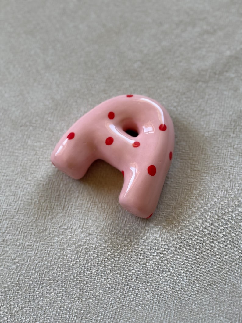Op de afbeelding: Een roze keramisch object met een gat in het midden en rode stippen. Het object heeft een gebogen vorm en is waarschijnlijk een decoratief item of een uniek kunstwerk.