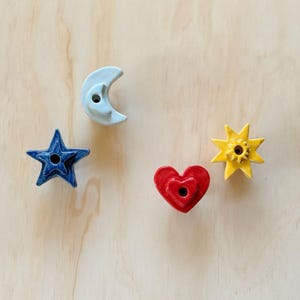 May include: Four colourful ceramic shapes: a blue star, a light blue crescent moon, a red heart, and a yellow sun. Each shape has a hole in the centre. The shapes are arranged on a light wood surface.