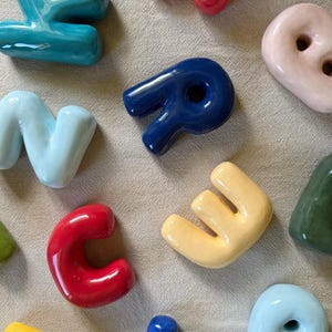 Op de afbeelding: Een verzameling kleurrijke keramische letters in verschillende tinten, waaronder rood, blauw, geel en groen. De letters zijn glanzend en lijken handgemaakt te zijn, met een glad, afgerond ontwerp. De letters zijn gerangschikt op een neutraal gekleurd stoffen oppervlak.