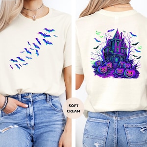 May include: Cream-colored t-shirt featuring a gradient bat design on the front and a Halloween haunted house graphic on the back. The back graphic includes a purple house, pumpkins, and bats. The shirt is labeled "Soft Cream".