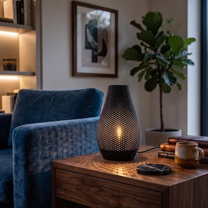Può includere: Una lampada da tavolo nera e strutturata con un bagliore caldo è appoggiata su un comodino in legno scuro. Il motivo della lampada proietta una luce a motivi sul tavolo. Sullo sfondo una poltrona blu in velluto, insieme a un quadro incorniciato e una pianta in vaso.
