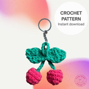 Puede incluir: Un llavero de cereza hecho a mano con un llavero plateado. El llavero presenta dos cerezas rosas y hojas verdes. El texto "CROCHET PATTERN Instant download" está en un círculo blanco. El logotipo de Ocean Friends Crochet Co. está en la esquina inferior derecha.