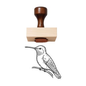 May include: A wooden rubber stamp featuring a dark brown handle and a light-colored rectangular wooden base. Below the stamp is a black and white line art illustration of a hummingbird perched on a thin branch, showing the stamp's imprint.