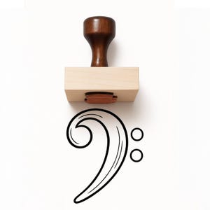 May include: A wooden rubber stamp featuring a dark brown handle and a light tan rectangular base. The stamp is positioned above a black ink impression of a bass clef musical symbol, which consists of a curved line and two small circles.
