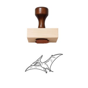 May include: A wooden rubber stamp featuring a dark brown handle and a light wood block base. Below the stamp is a black and white line art illustration of a pterodactyl in flight with its wings spread wide, showing the design it creates.