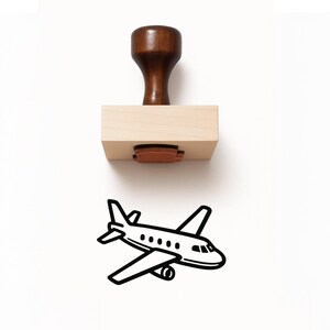 May include: A wooden stamp with a dark brown handle and a light-colored rectangular base. The stamp's design is a black outline of an airplane. The stamp is ideal for adding a travel theme to paper crafts or stationery.