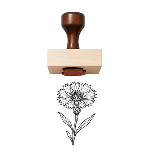 May include: A wooden rubber stamp featuring a dark brown handle and a light tan rectangular base. Below the stamp is a black and white line art illustration of a single cornflower, showing the detailed petals, a textured center, and leaves.