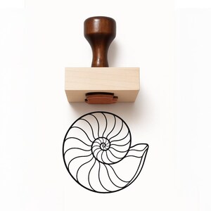 May include: A wooden rubber stamp featuring a dark brown handle and a light wood block base. Below the stamp is a black ink impression of a nautilus shell, displaying a detailed spiral pattern with curved lines radiating from the center point.