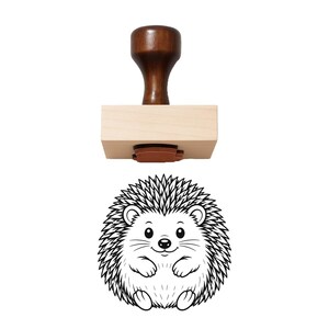 May include: A wooden rubber stamp featuring a dark brown handle and a light wood base. Below the stamp is a black and white line art illustration of a round, smiling hedgehog with detailed quills, small ears, and paws, centered on a white background.