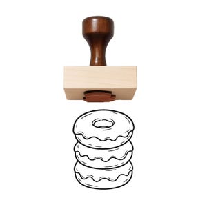 May include: A wooden rubber stamp featuring a dark brown handle and a light wood base. The stamp die is made of orange rubber and displays a black line art illustration of a stack of three glazed donuts with icing drips on each one.