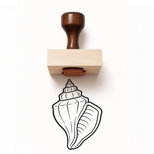 May include: A wooden rubber stamp featuring a dark brown handle and a light-colored rectangular wooden base. Below the stamp, a black ink impression shows a detailed line drawing of a spiral seashell with a textured interior and pointed top.