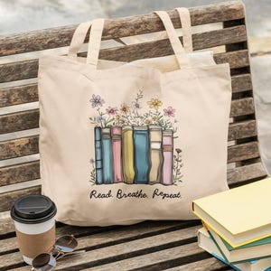 以下が含まれることがあります： 本と花のカラフルなイラストが描かれたベージュのトートバッグ。「Read. Breathe. Repeat.」の文字が下にプリントされています。コーヒーカップとサングラスが木製のベンチに置かれています。