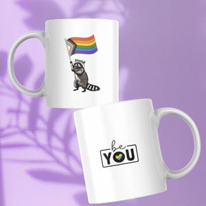 May include: Two white ceramic mugs. One mug features a raccoon holding a rainbow flag. The other mug has the words "be YOU" with a rainbow heart. Both mugs have a white handle and a purple background.