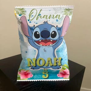 以下が含まれることがあります： リロ＆スティッチのキャラクター、スティッチが描かれた青いパーティーバッグ。バッグの上部には緑色の筆記体で「Ohana」の文字。名前「Noah」と数字「5」も表示されています。ピンク色のハイビスカスの花が底を飾っています。