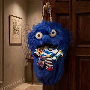 May include: A whimsical blue monster-shaped sock holder with large googly eyes, hanging from a door. The holder is filled with colorful socks of various patterns and designs. The plush material and playful design add a fun element to any room.