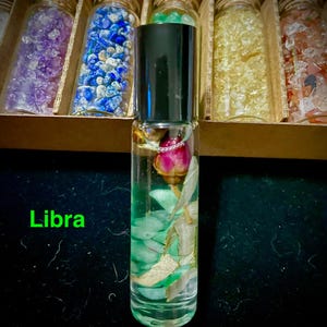 May include: A clear glass roll-on bottle with a black cap, containing a pink rose bud, green stones, and gold flakes. The word "Libra" is written in green. The bottle is in front of a wooden box with several glass jars filled with colorful stones.