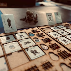 Può includere: Un'impostazione di gioco da tavolo con pezzi e carte in legno. Il gioco include carte con illustrazioni di abbigliamento e altri oggetti. Una carta con il testo "Rashide Arms" è visibile, insieme a una miniatura.