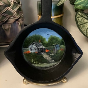 May include: A black cast iron skillet with a painted scene of a white house with a green roof and a blue sky. The house is surrounded by trees and flowers. The skillet is on a gold stand.