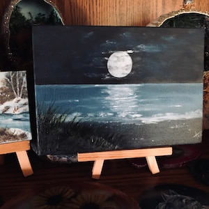 May include: A small painting on canvas depicting a moonlit seascape. The painting is on a wooden easel and features a full moon reflecting on the water.