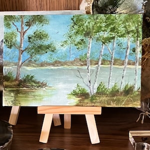 May include: A small painting on canvas depicting a lake scene with trees and a blue sky. The painting is on a wooden easel.