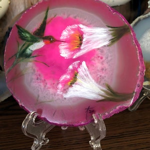 May include: A hand-painted agate coaster featuring a hummingbird and two white flowers on a pink background. The coaster is on a clear plastic stand. The artist's signature is visible in the lower right corner.