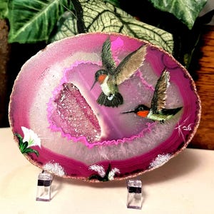May include: An oval-shaped pink agate slice featuring a hand-painted scene of two hummingbirds with orange throats in flight. The center of the stone displays a textured, crystalline geode cavity. White flowers are painted at the bottom and side.