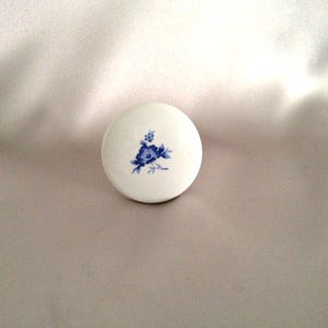 May include: A round, white ceramic cabinet knob featuring a small, centered blue floral pattern. The knob has a smooth, glossy finish and a convex shape, displaying a delicate botanical illustration in shades of blue against the white surface.