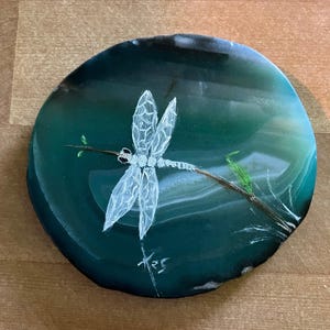 May include: A round, polished agate stone coaster with a hand-painted dragonfly design. The dragonfly is white with delicate wings, perched on a brown branch with green leaves. The agate has shades of green and white.