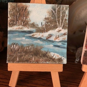 May include: A small painting on a wooden easel depicts a winter scene with a blue river, brown trees, and white snow. The painting is on a canvas and is approximately 4 inches by 4 inches.