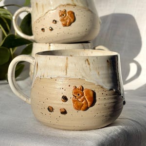 Könnte beinhalten: Zwei handgefertigte Keramiktassen mit beiger, gesprenkelter Basis und weiß glasierter Oberseite. Jede Tasse zeigt ein erhabenes, orange-braunes Eichhörnchen-Relief und kleine dunkelbraune Eichel-Akzente an der Seite. Abgerundete Form mit Henkel.