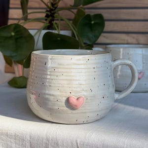 Può includere: Una tazza in ceramica rotonda color bianco sporco con finitura maculata e striature orizzontali. Un piccolo cuore in rilievo rosa chiaro è centrato sulla parte anteriore. La tazza presenta un manico curvo sul lato destro, una seconda tazza è visibile.