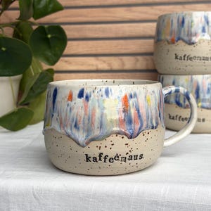 Könnte beinhalten: Ein Keramikbecher mit beigefarbener, gesprenkelter Basis und einer farbenfrohen Tropfglasur in Blau-, Orange- und Gelbtönen am oberen Rand. Das Wort kaffeemaus ist in schwarzen Kleinbuchstaben auf der Vorderseite aufgedruckt. Ein Henkel ist rechts.