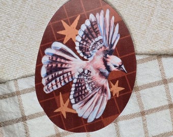 May include: An oval sticker with a watercolour illustration of a blue jay in flight. The bird has pink, blue, and white feathers, set against a brown background with orange star accents. The sticker is placed on a beige and white checkered fabric.