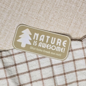 May include: A rectangular sticker with a tan background and white text that reads "NATURE IS AWESOME!" with a tree graphic. Below the text is the phrase "(But ticks freak me out)". The sticker is on a beige and brown checkered surface.