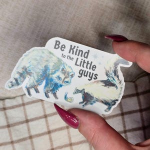 May include: A sticker featuring a raccoon and a skunk with the text "Be Kind to the Little Guys." The raccoon is blue and white, and the skunk is black and white. A small snail is also depicted.
