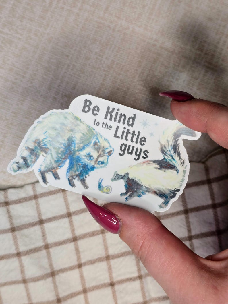 May include: A sticker featuring a raccoon and a skunk with the text "Be Kind to the Little Guys." The raccoon is blue and white, and the skunk is black and white. A small snail is also depicted.