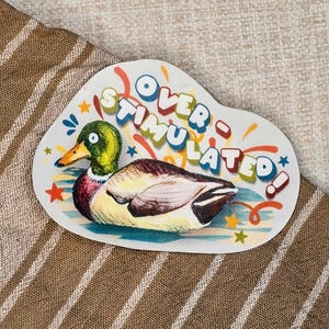 May include: A white, irregular-shaped sticker featuring a cartoon duck with the text "OVER STIMULATED!" in colorful letters. The duck has a green head, yellow beak, and brown and white body. Stars and decorative elements surround the text.