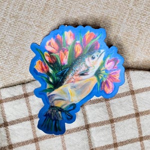 May include: A sticker featuring a fish nestled among a bouquet of tulips. The fish is detailed with scales and fins, while the tulips display shades of red, orange, and pink. The bouquet is wrapped in a paper cone and tied with a blue ribbon, all set against a blue border.