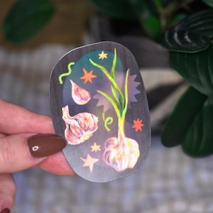 May include: A die-cut sticker with a dark background featuring illustrations of garlic bulbs and green scallions. The design includes small orange and yellow star shapes, a green squiggly line, and a purple-toned glow effect behind the vegetables.