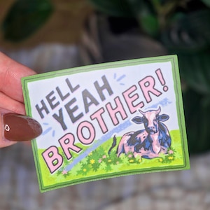 May include: A rectangular sticker featuring a black and white cow resting in a green field with yellow flowers. The text HELL YEAH BROTHER! is printed in large pink letters across the top left, accented by small light blue lines.