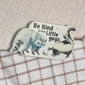 May include: A rectangular sticker featuring a raccoon and a skunk, with the text "Be Kind to the Little Guys." The raccoon is depicted in shades of blue and gray, while the skunk is black and white. A small snail is also present.